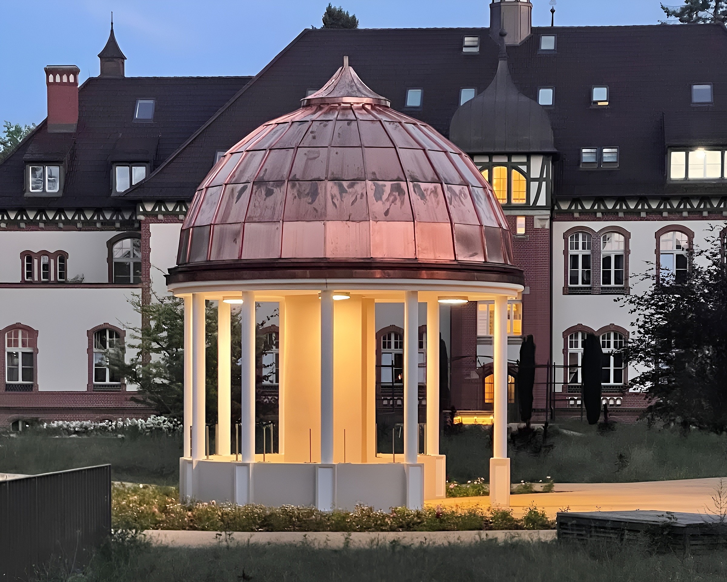 Ein runder Pavillon mit glänzender Kupferkuppel und weißen Säulen ist in der Abenddämmerung warm beleuchtet. Dahinter erhebt sich ein historisches Gebäude mit dunklem Schieferdach, Fachwerkgiebeln, Backsteinrahmen um die Fenster und einem Zwiebelturm. Vor dem Pavillon wachsen niedrige Sträucher und Rasen.