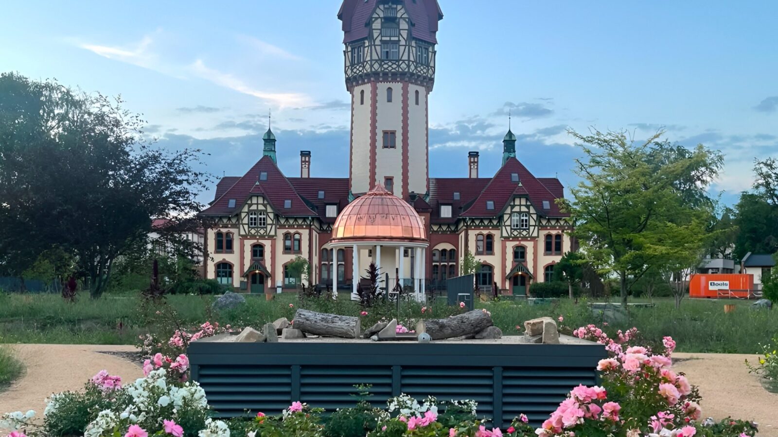 Ein historisches Gebäude mit hohem, spitz zulaufendem Turm im Fachwerk- und Backsteinstil steht unter blauem Abendhimmel. Vor dem Haupteingang leuchtet eine kupferne Kuppel auf weißen Säulen. Im Vordergrund blühen rosafarbene und weiße Rosen in einem Beet mit Rindenmulch, dahinter ein dunkler, lamellenverkleideter Lüftungsschacht mit Holzstämmen auf dem Deckel.